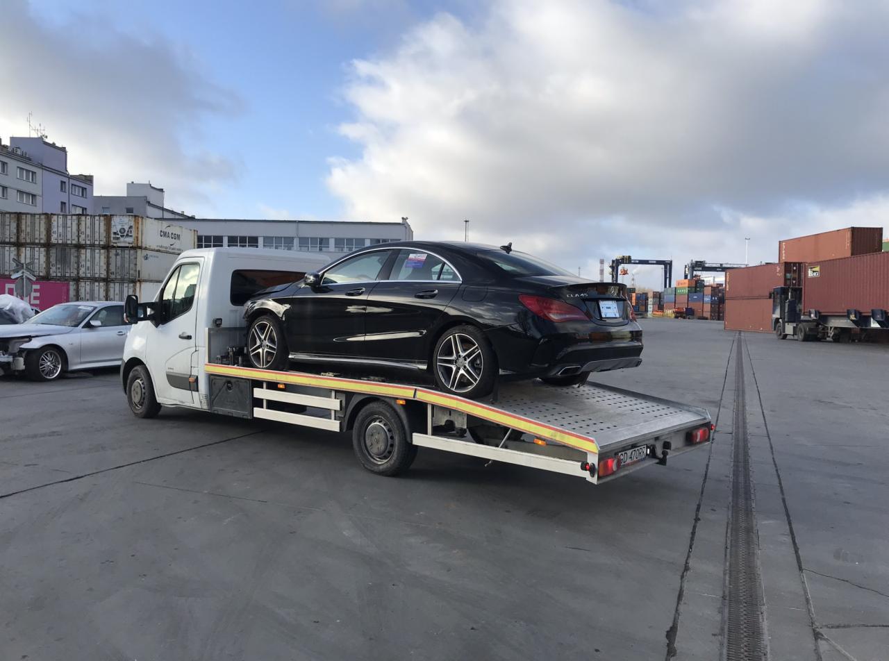 Czarny Mercedes CLA na platformie lawety w porcie kontenerowym, widoczny uszkodzony srebrny samochód w tle.