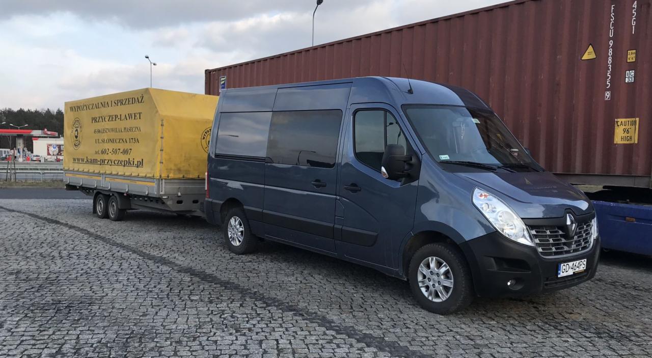 Niebieski bus Renault Master ciągnie przyczepę-lawetę z żółtą plandeką, na której widnieje napis 'WYPOŻYCZALNIA I SPRZEDAŻ PRZYCZEP-LAWET' oraz dane kontaktowe firmy, na tle parkingu z kostki...
