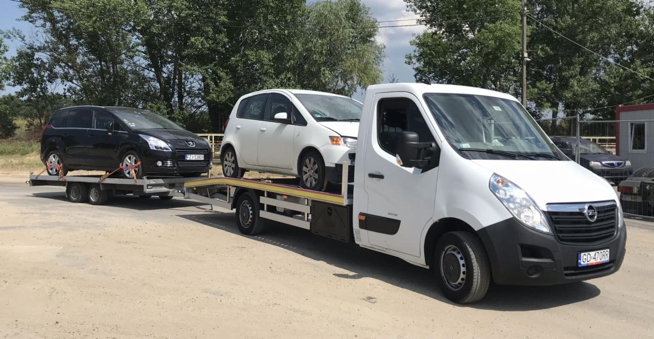 Biała laweta z dwoma samochodami osobowymi: czarnym Peugeotem i białym Mitsubishi, zabezpieczonymi pasami transportowymi, na tle drzew i budynków.