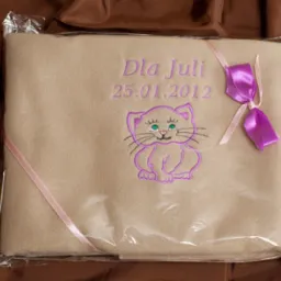 Beżowy kocyk zapakowany w folię, z fioletowym haftem przedstawiającym kota, imieniem 'Dla Juli' i datą 25.01.2012, przewiązany różową i fioletową wstążką.