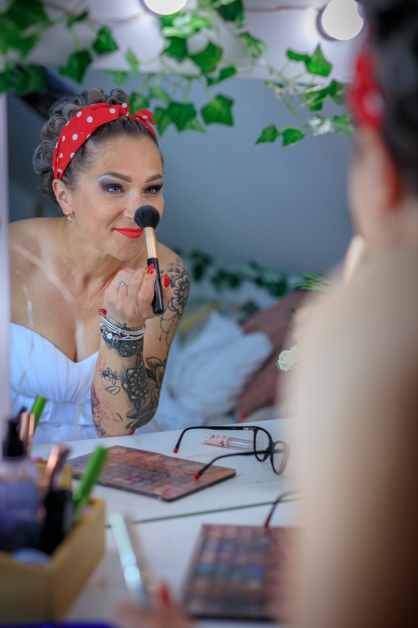 Kobieta z czerwoną opaską w białe kropki, z tatuażami na ramieniu, aplikuje makijaż pędzlem przed lustrem, otoczonym girlandą z zielonych liści.