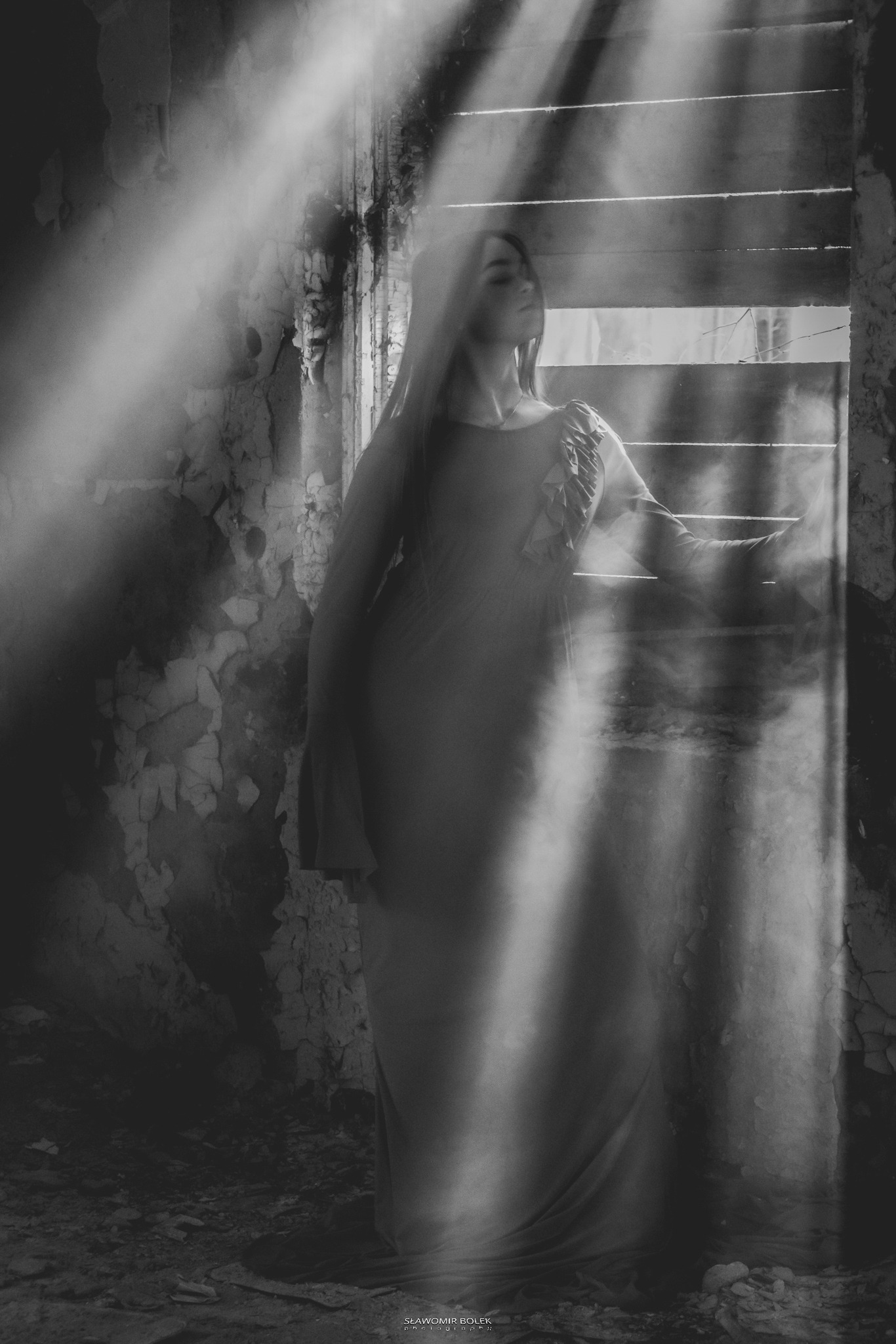 Kobieta w długiej sukni pozuje w zrujnowanym wnętrzu, promienie światła wpadają przez okno z żaluzjami, czarno-biała fotografia.