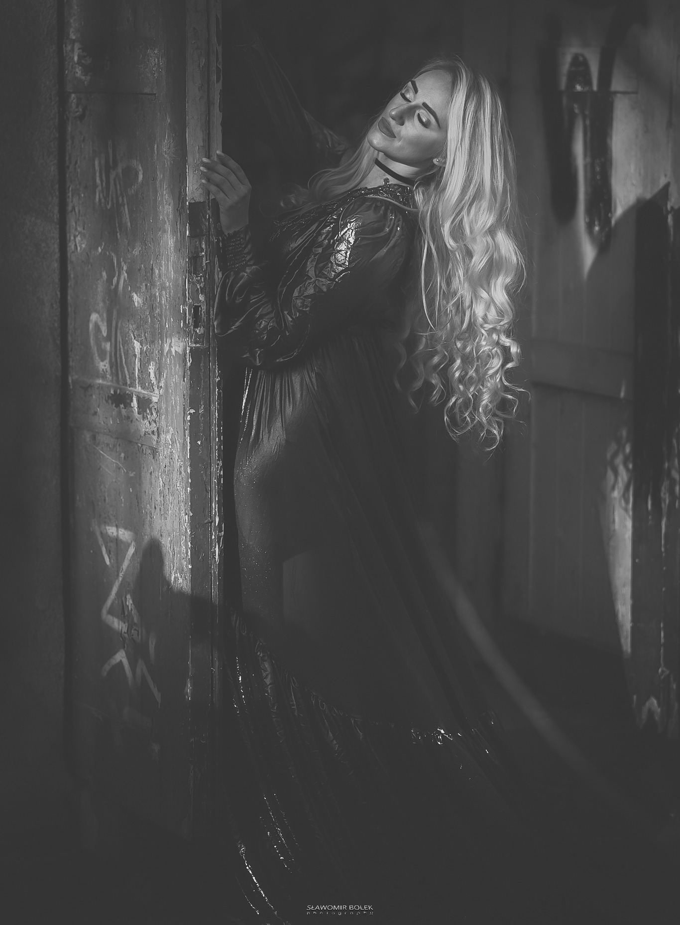 Portret kobiety w długiej sukni, opierającej się o starą, pomazaną ścianę, w ciemnym otoczeniu, z długimi blond włosami i zamkniętymi oczami.