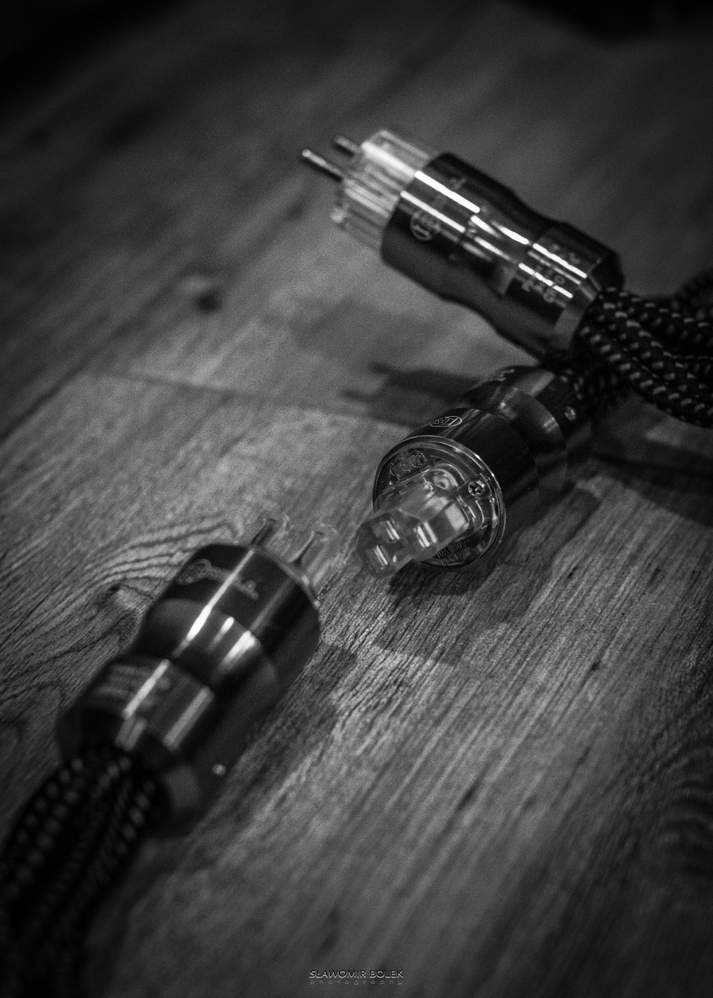 Czarno-białe zbliżenie trzech wtyczek z przezroczystymi elementami i plecionymi kablami na drewnianym tle, ukazujące detale konstrukcji i fakturę materiałów.