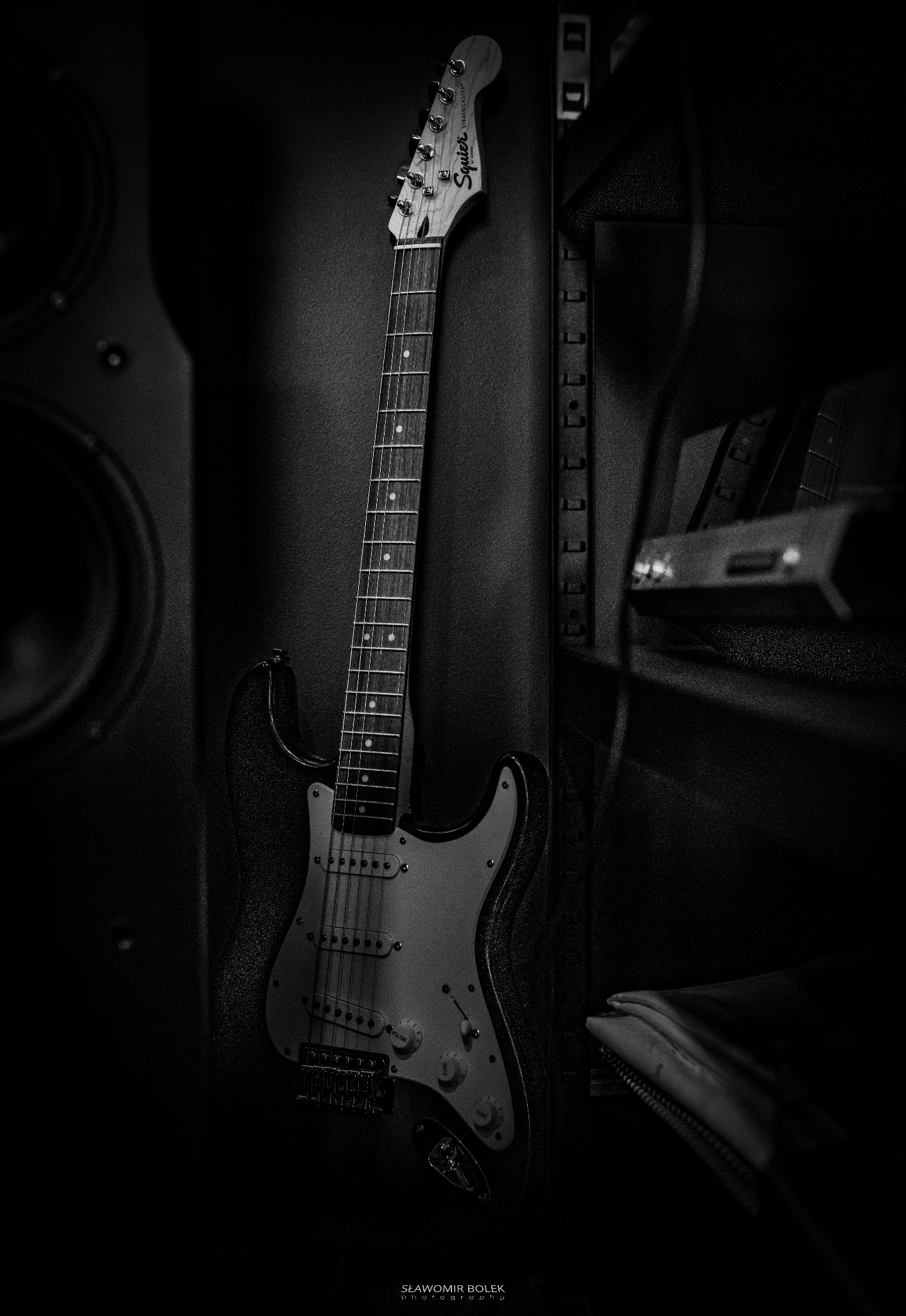 Czarno-białe zdjęcie gitary elektrycznej Squier Stratocaster oparta o wzmacniacz, z widocznym logo na główce i elementami panelu kontrolnego