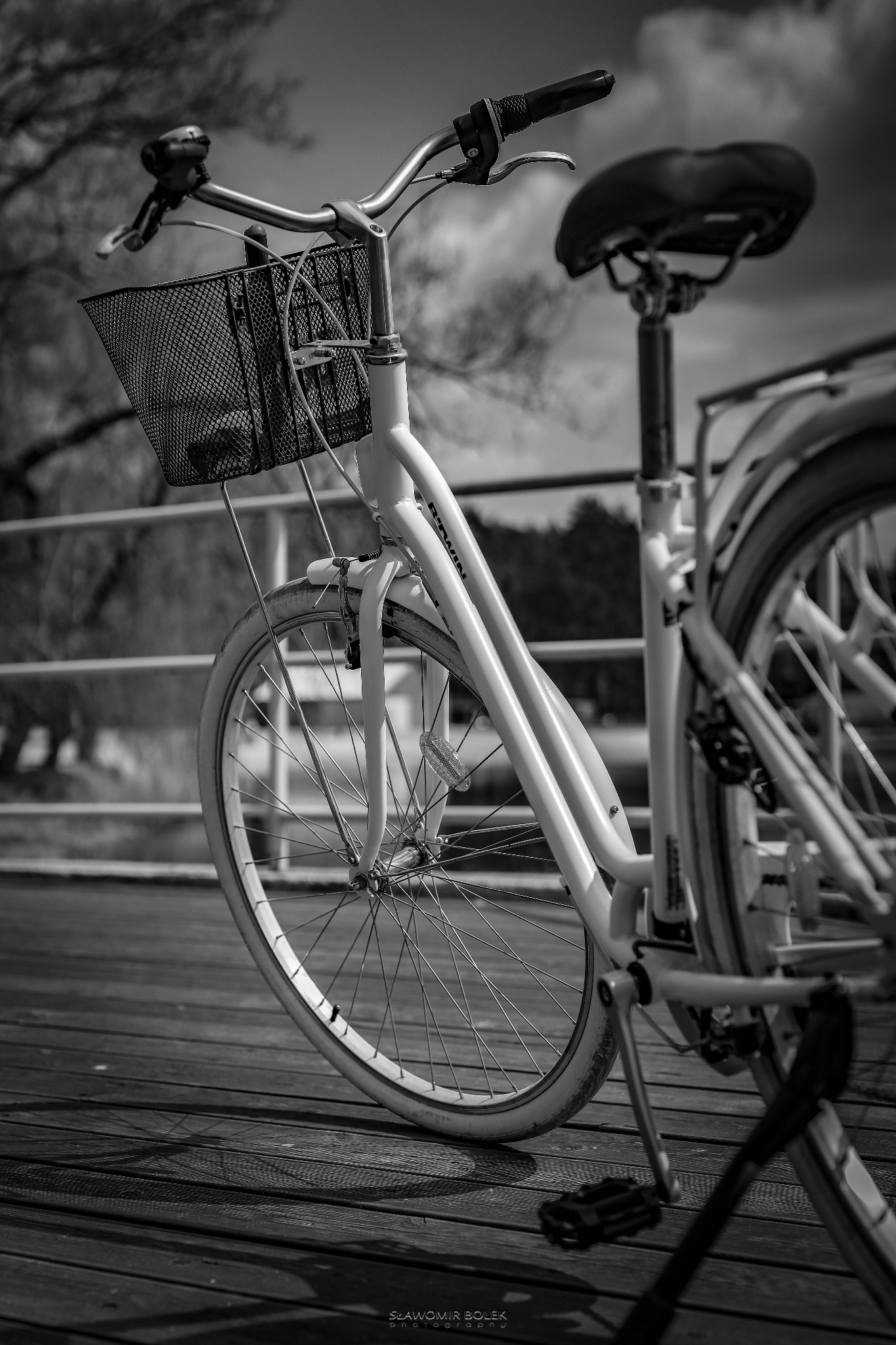 Czarno-białe zdjęcie roweru miejskiego z koszykiem na drewnianym pomoście, widok z bliska na przednie koło i kierownicę, z fragmentem siodełka w tle.