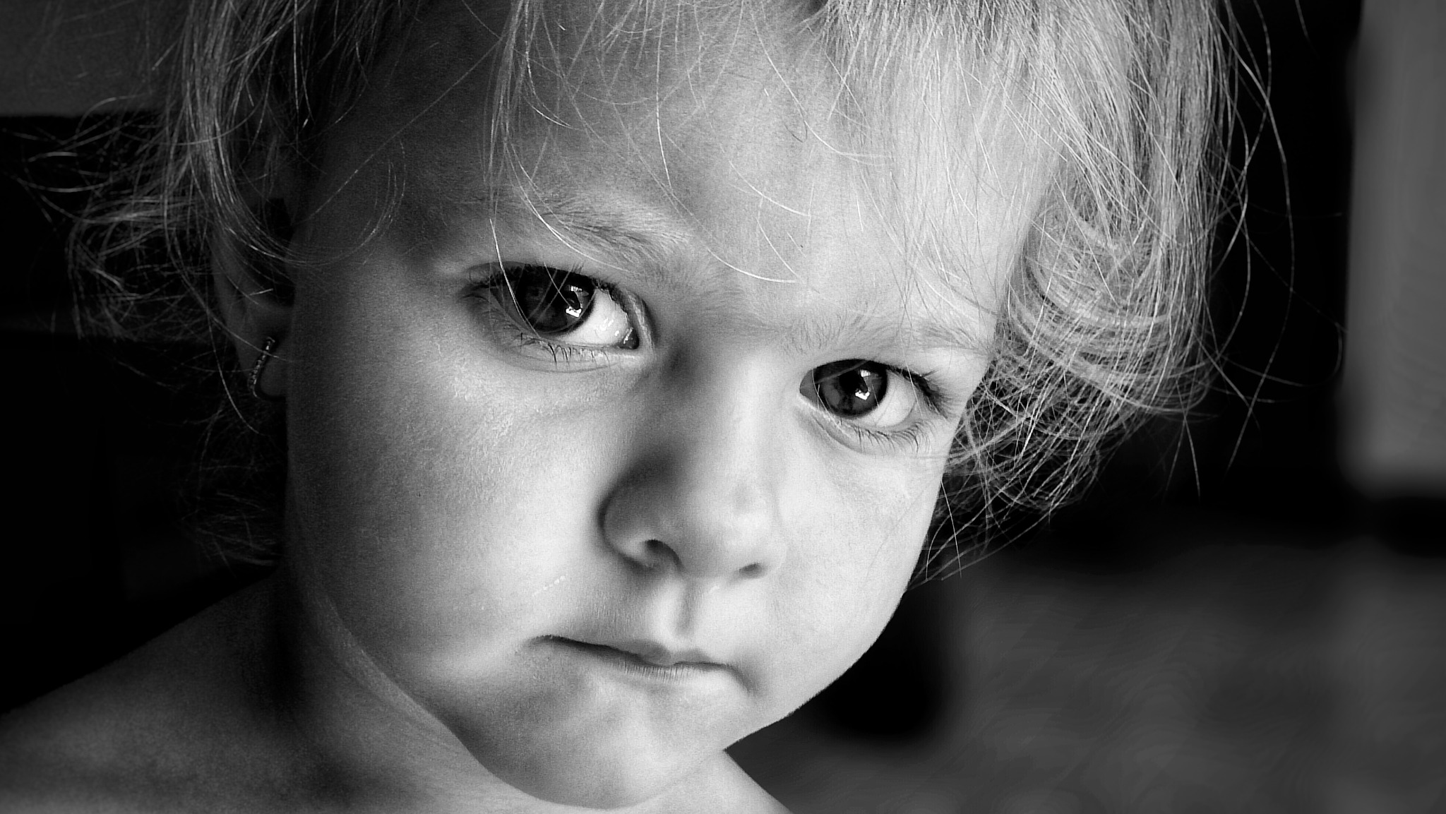 Portret małej dziewczynki z poważnym wyrazem twarzy, czarno-białe zdjęcie z bliska, widoczne delikatne włosy i kolczyk w uchu.