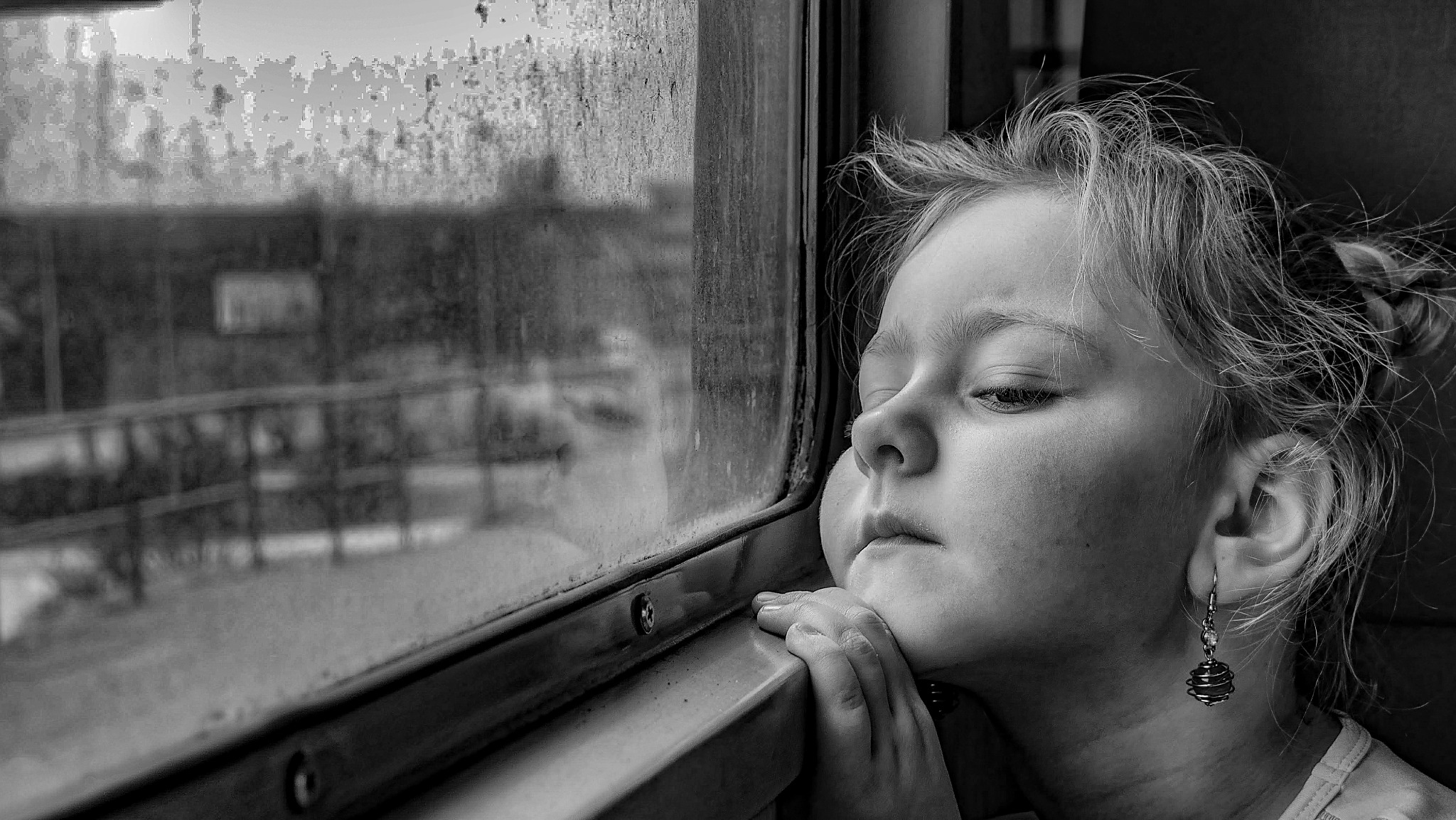 Zamyślona dziewczynka opiera brodę na dłoniach, spoglądając przez brudną szybę pociągu, w czerni i bieli, z widocznym kolczykiem w uchu.