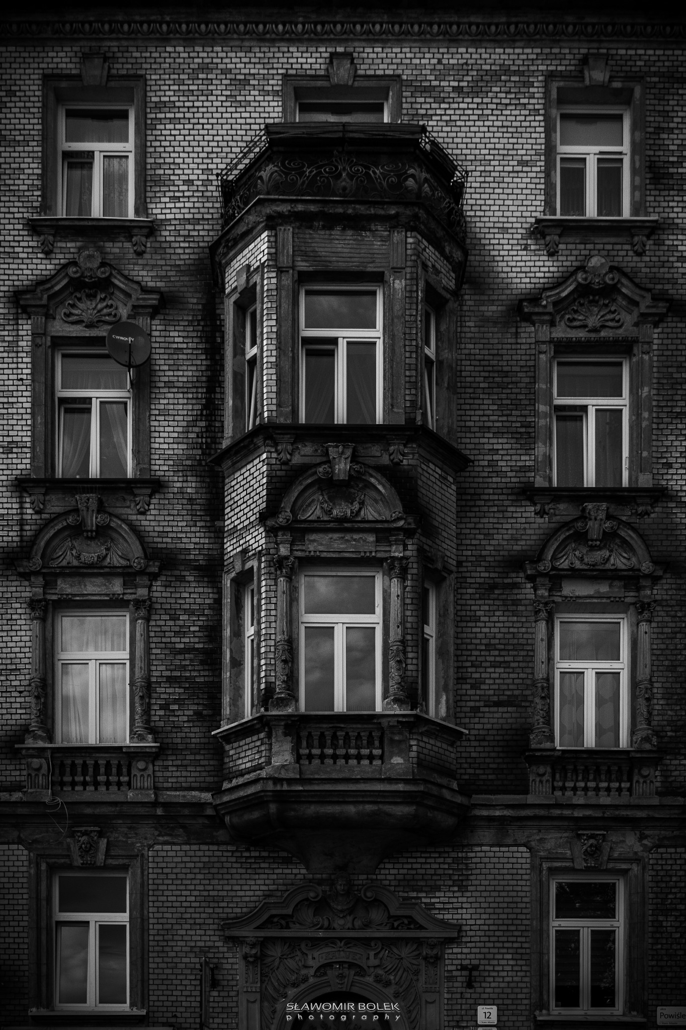 Czarno-białe zdjęcie fasady starej kamienicy z cegły z wyraźnym, wystającym balkonem i ozdobnymi ramami okiennymi, na jednym z okien widoczna antena satelitarna.