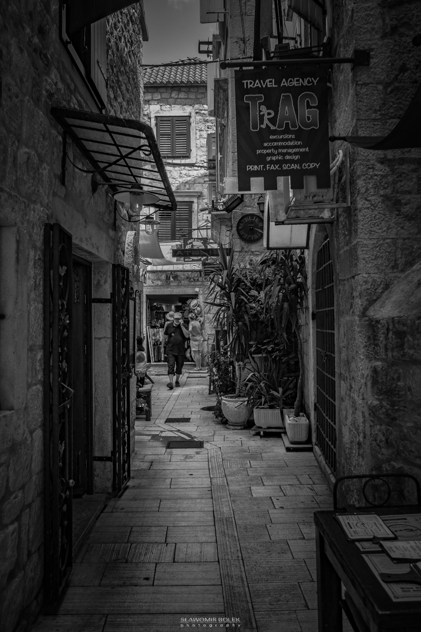 Czarno-białe zdjęcie wąskiej uliczki w starym mieście z kamiennymi budynkami, roślinami w donicach, szyldem agencji turystycznej 'TRAG' i przechodzącymi ludźmi w oddali.