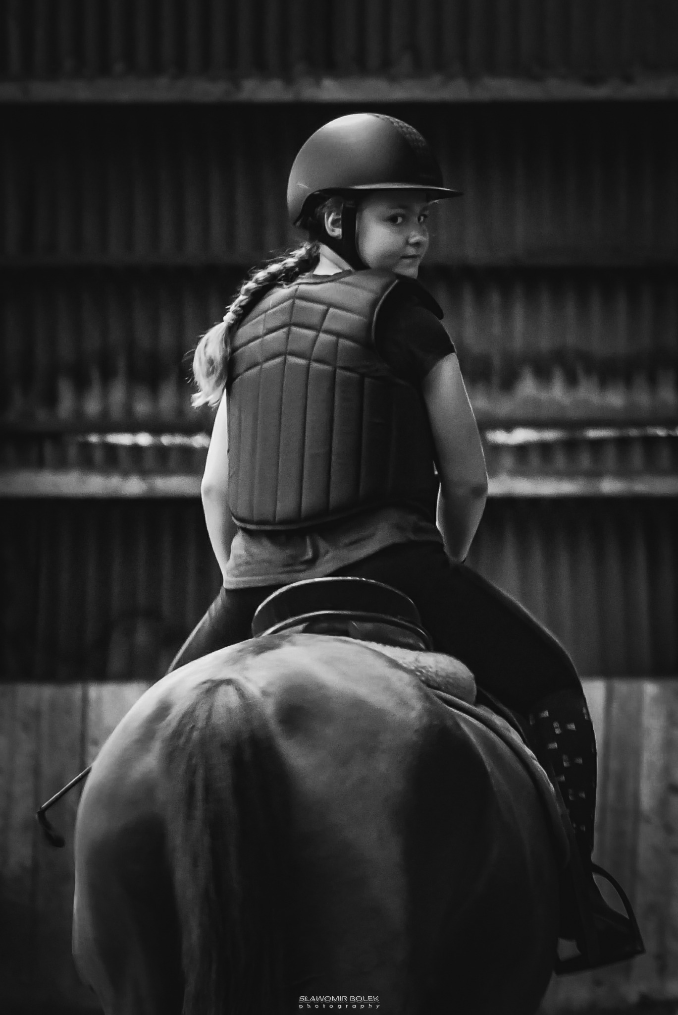 Czarno-białe zdjęcie dziewczynki w kasku i kamizelce ochronnej siedzącej na koniu, z warkoczem opadającym na plecy, spoglądającej przez ramię w stajni.