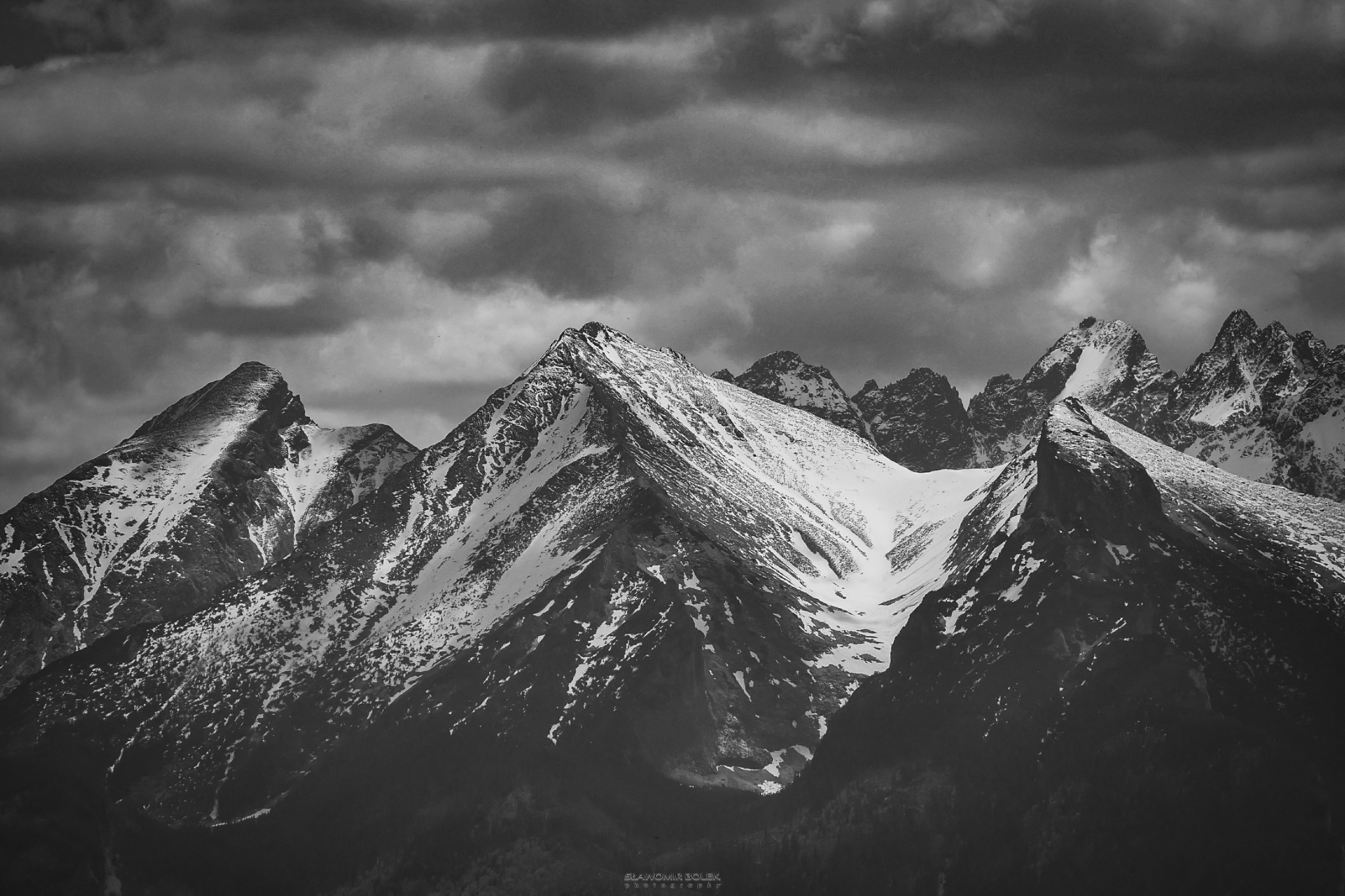 Czarno-biała fotografia górskich szczytów pokrytych śniegiem, z dramatycznym, pochmurnym niebem w tle.