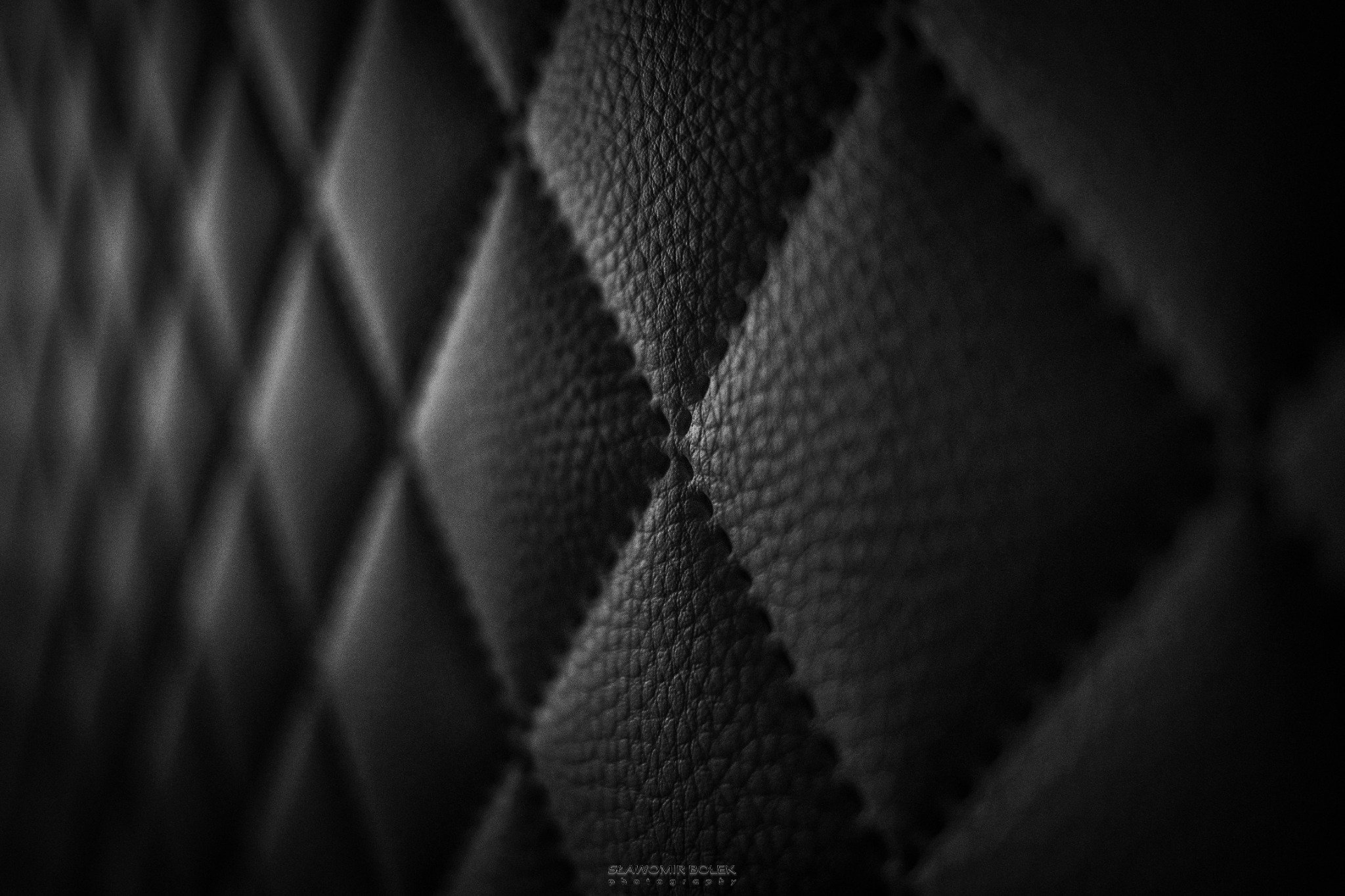 Czarno-białe zbliżenie na pikowaną, skórzaną tapicerkę z wyraźnym szwem w kształcie rombów, oświetloną z boku, z delikatnym efektem rozmycia w tle.