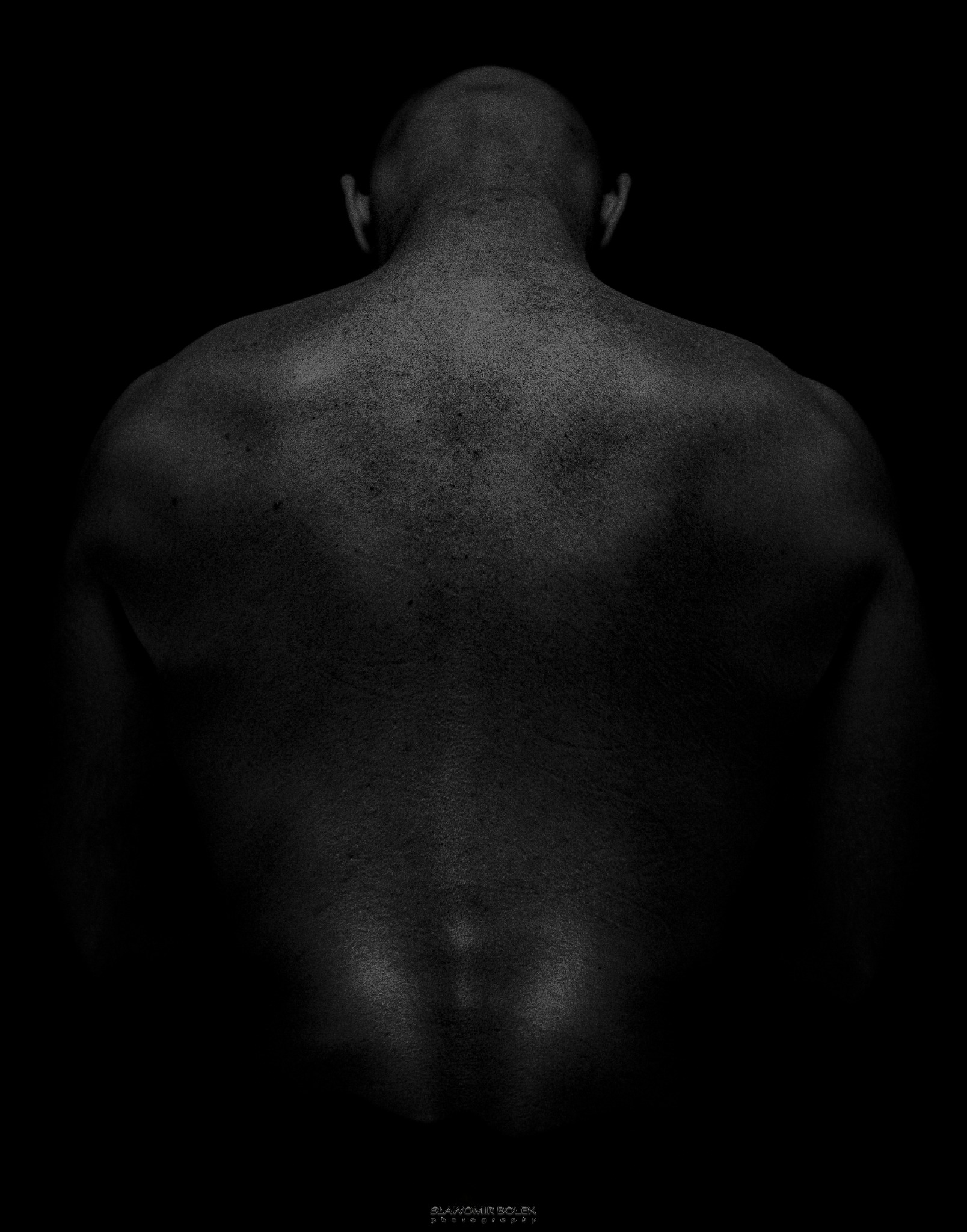 Czarno-białe zdjęcie pleców i karku mężczyzny na ciemnym tle, widoczne detale skóry i delikatne światłocienie.