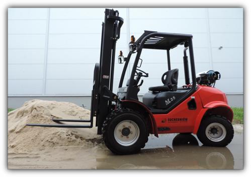 Czerwony wózek widłowy M25 marki Suchodniołów z czarnym masztem i widłami, stojący na mokrym placu obok pryzmy piasku na tle szarej hali.