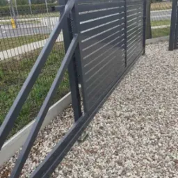 Nowoczesna, przesuwna brama w kolorze grafitowym z poziomymi panelami, zamontowana na żwirowym podłożu, widoczna z boku w częściowym otwarciu.