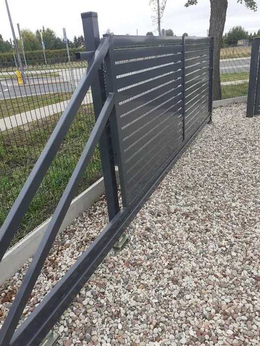 Nowoczesna, przesuwna brama w kolorze grafitowym z poziomymi panelami, zamontowana na żwirowym podłożu, widoczna z boku w częściowym otwarciu.