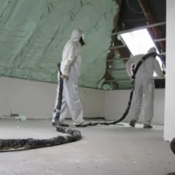 Pracownicy w kombinezonach ochronnych aplikują pianę izolacyjną na poddaszu, widoczny wąż i pokryta pianą skośna ściana poddasza.