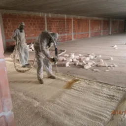 Pracownicy w kombinezonach ochronnych natryskują pianę izolacyjną na betonową powierzchnię wewnątrz budynku z ceglanymi ścianami, widoczne bloki styropianu.