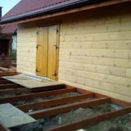 Drewniany budynek gospodarczy z brązowym dachem w trakcie budowy tarasu: widoczne drewniane legary tworzące konstrukcję pod taras, ułożone na ziemi z częściowo położoną płytą tymczasową.