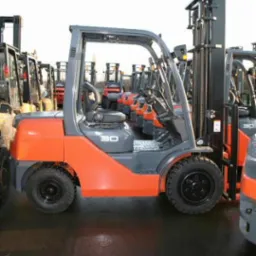 Pomarańczowo-szary wózek widłowy Toyota, model 30, stoi w rzędzie z innymi wózkami. Widok z boku, opony z bieżnikiem, czarne siedzenie operatora.