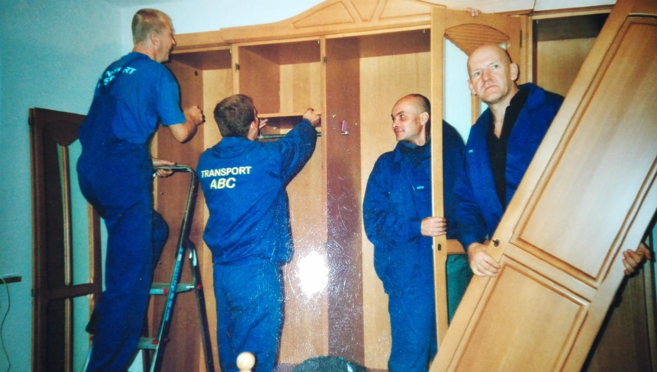 Czterej pracownicy w niebieskich kombinezonach firmy Transport ABC montują dużą, drewnianą szafę w pomieszczeniu podczas relokacji.