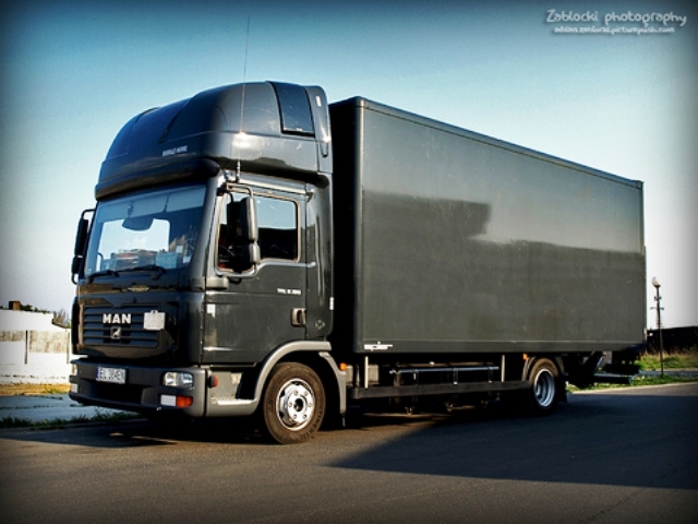 Szary samochód ciężarowy MAN z zabudową kontenerową, zaparkowany na asfaltowej drodze w słoneczny dzień.