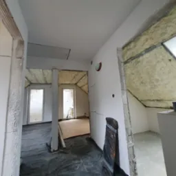 Poddasze w trakcie remontu: widoczna izolacja termiczna na skośnych ścianach, surowy stan ścianek działowych z widocznymi pęknięciami, zabezpieczona folią podłoga i fragment okna dachowego.