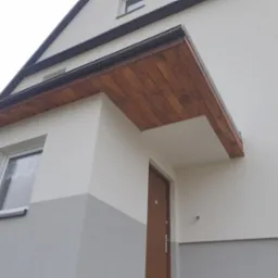 Wykończona elewacja domu z drewnianym zadaszeniem nad wejściem i oknem, ściany pomalowane w dwóch odcieniach szarości i bieli, widok z dołu.
