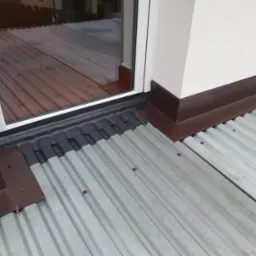 Szczegółowe ujęcie obróbki blacharskiej przy oknie dachowym z widoczną membraną dachową i blachą trapezową, brązowe elementy wykończeniowe.