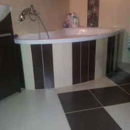 Wykończona łazienka z narożną wanną obudowaną brązowo-beżowymi płytkami. Na podłodze płytki w szachownicę. Umywalka z szafką po lewej stronie.