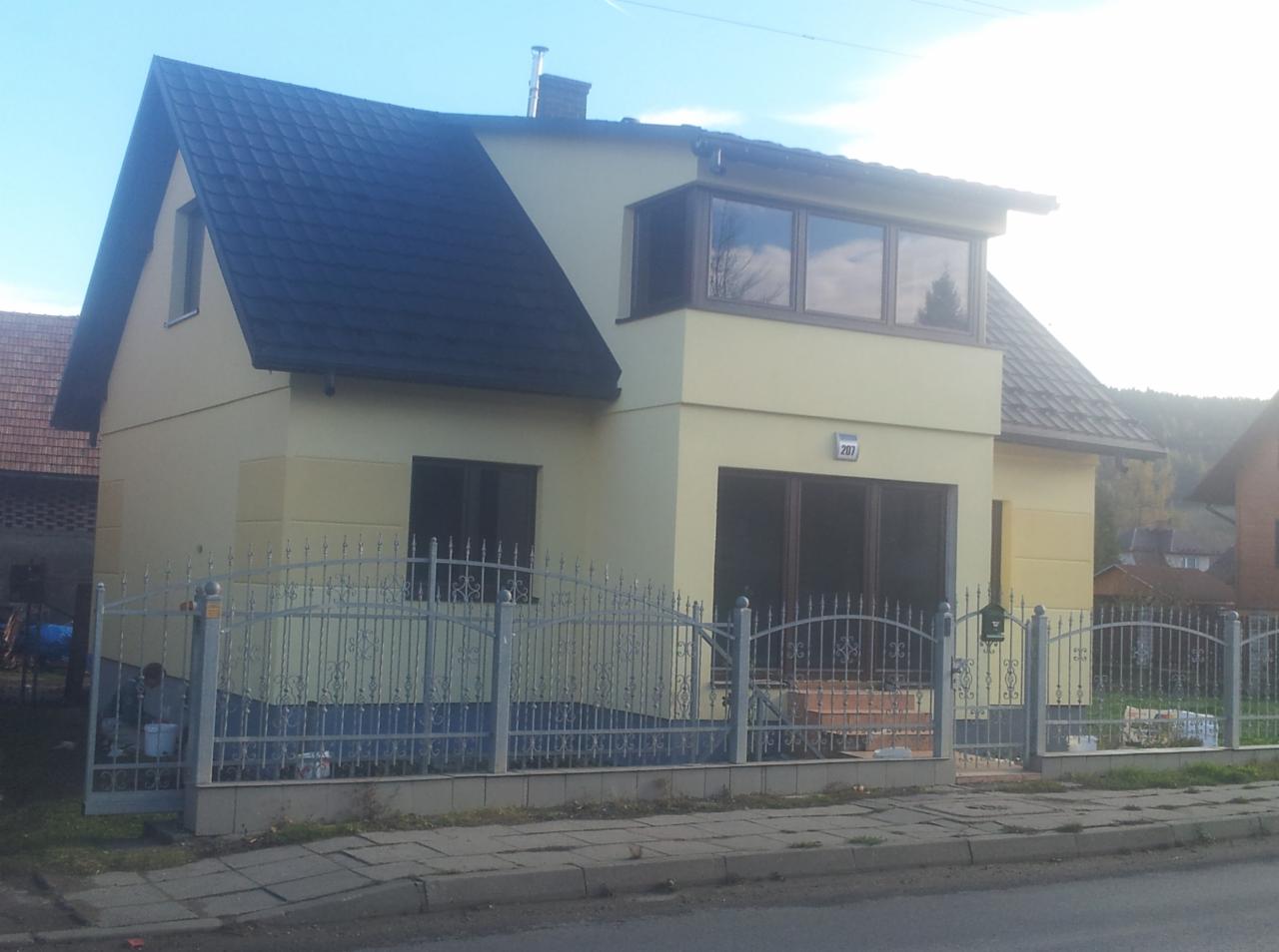 Żółty dom jednorodzinny z ciemnym dachem, szarą metalową bramą i zieloną skrzynką na listy. Widoczny fragment elewacji sąsiedniego domu i drzewa w tle.