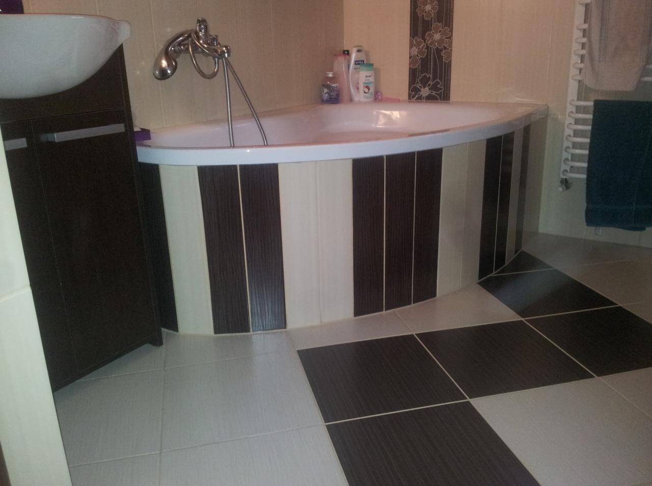 Wykończona łazienka z narożną wanną obudowaną brązowo-beżowymi płytkami. Na podłodze płytki w szachownicę. Umywalka z szafką po lewej stronie.