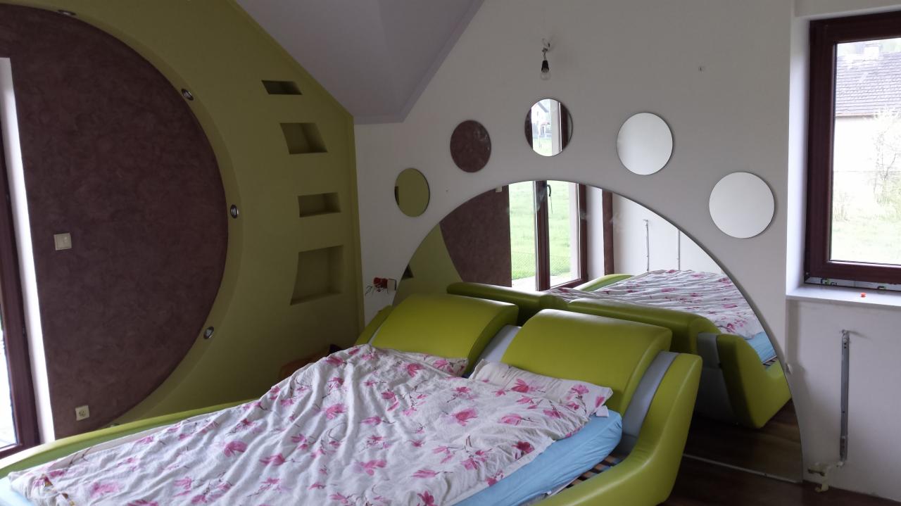Awangardowa sypialnia z łóżkiem w odcieniach zieleni, okrągłymi lustrami i dekoracyjną ścianą w kształcie półkola. Nietypowe oświetlenie i okno z brązową ramą.