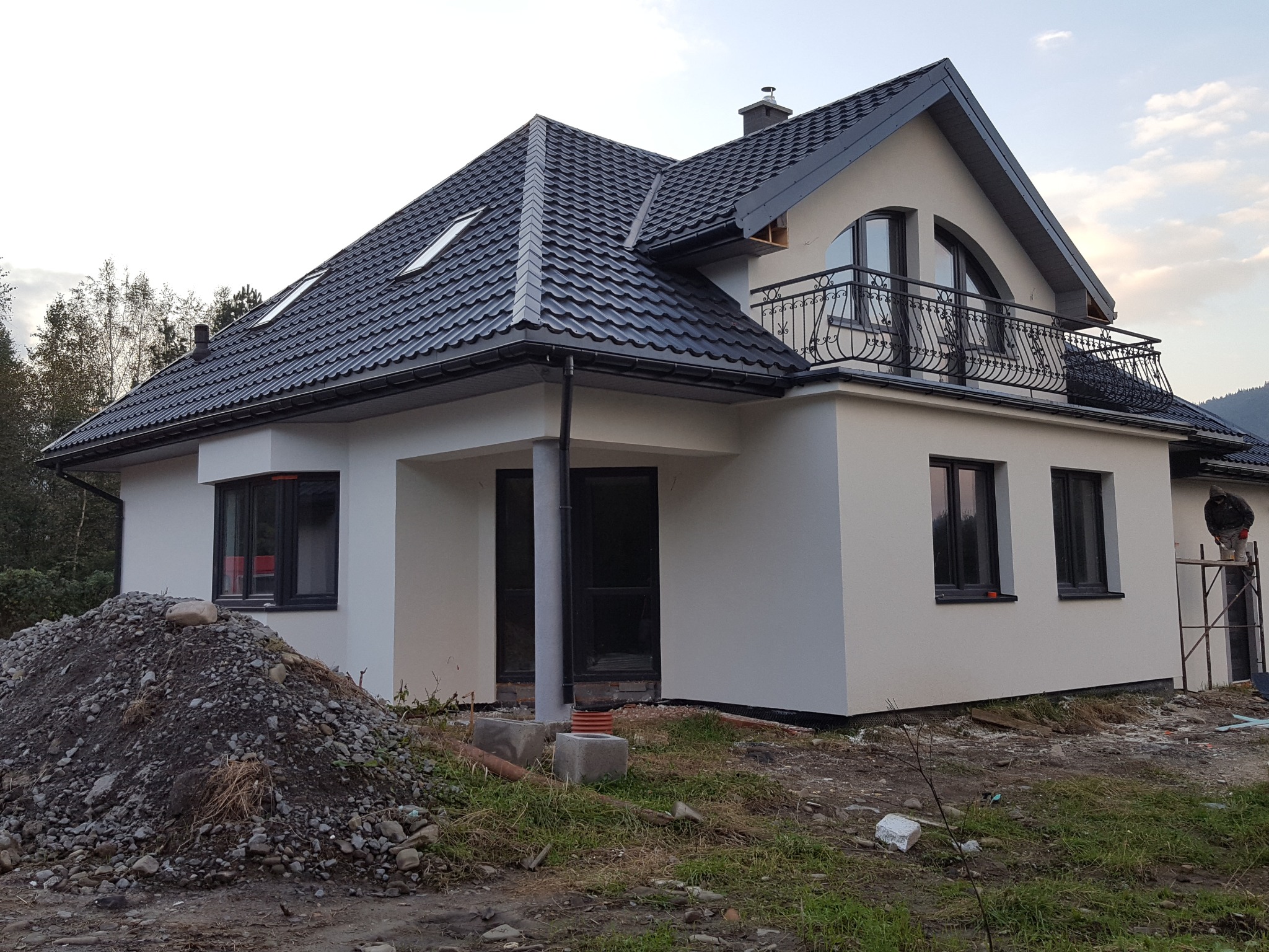 Nowoczesny dom jednorodzinny z szarą elewacją i ciemną dachówką, widoczny fragment elewacji w trakcie prac wykończeniowych, z rusztowaniem i stertą ziemi przed budynkiem.