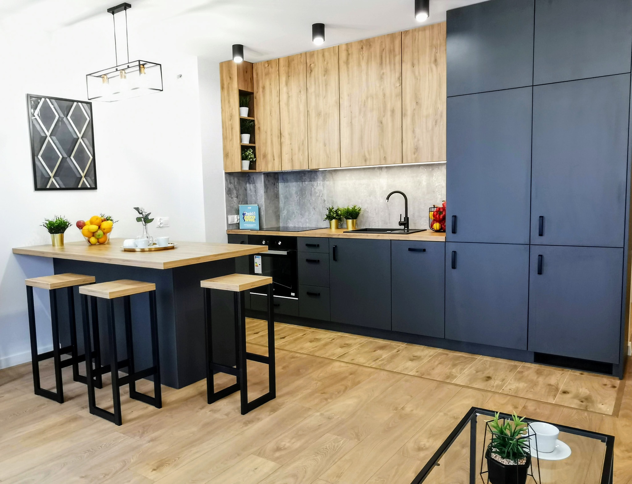 Nowoczesna kuchnia z drewnianymi i grafitowymi szafkami, wyspą z hokerami, oświetleniem punktowym i geometrycznym obrazem na ścianie.