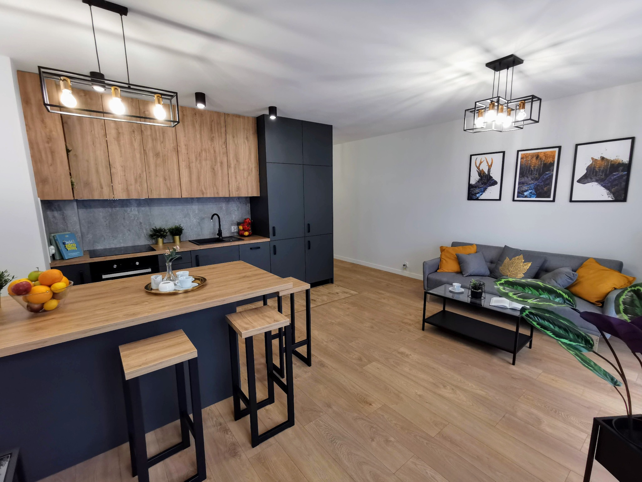 Nowoczesne wnętrze: otwarta kuchnia z wyspą i hokerami, szare szafki, drewniane blaty, salon z szarą sofą, obrazami zwierząt i roślinami doniczkowymi, oświetlenie w stylu industrialnym.