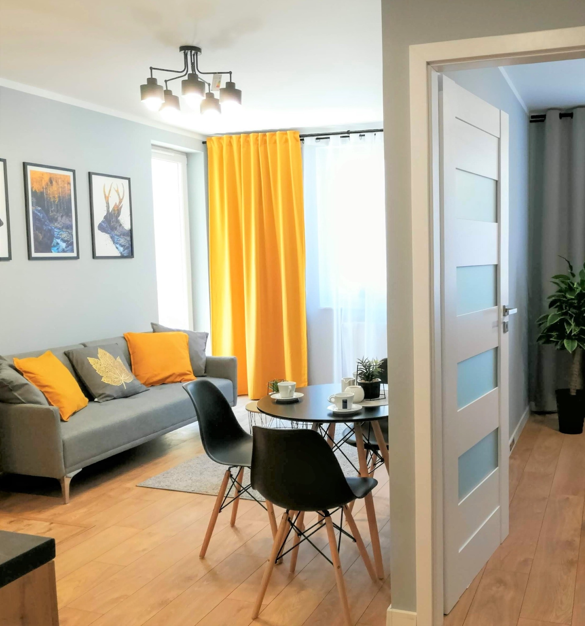 Jasne wnętrze pokoju dziennego z szarą sofą z żółtymi poduszkami, okrągłym stolikiem kawowym i czarnymi krzesłami na drewnianej podłodze, akcentowane żółtymi zasłonami i białymi drzwiami...