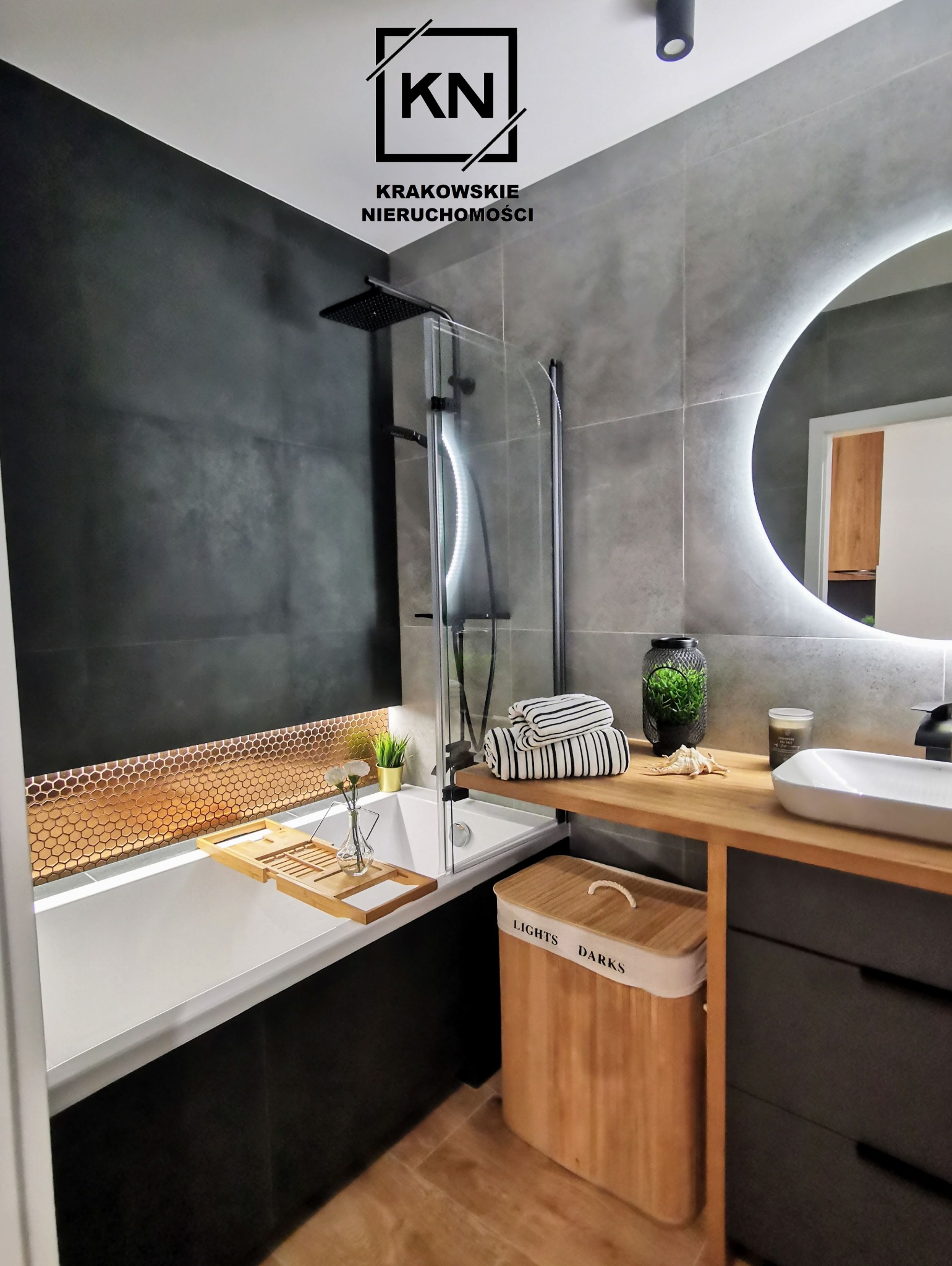 Nowoczesna łazienka z czarną armaturą, mozaiką w kolorze miedzi nad wanną i okrągłym lustrem z podświetleniem LED. Widoczna bambusowa taca na wannie i kosz na pranie z napisem 'Lights Darks'.