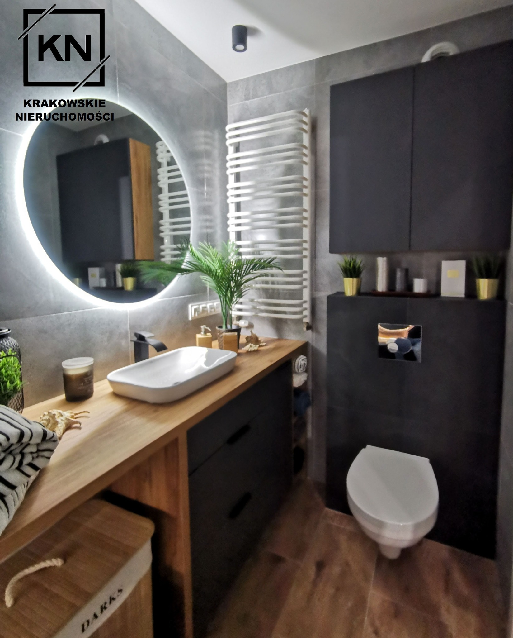 Nowoczesna łazienka z szarymi płytkami, drewnianym blatem pod umywalką, okrągłym lustrem z podświetleniem LED, czarnymi szafkami i wiszącą toaletą.