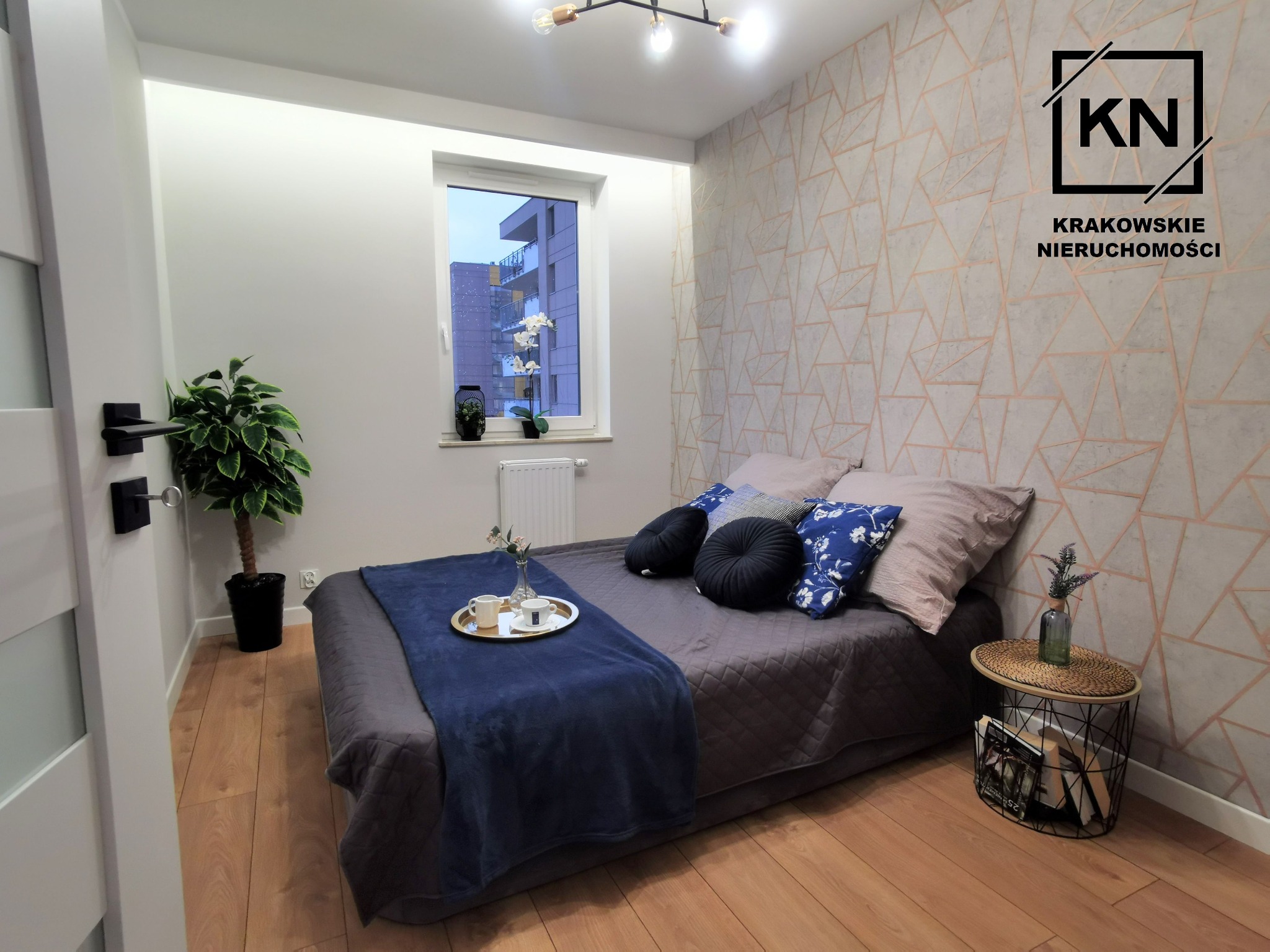 Nowoczesna sypialnia z łóżkiem, stolikiem nocnym z wiklinowym blatem i geometryczną tapetą w odcieniach beżu i szarości, widok na fragment budynku za oknem.