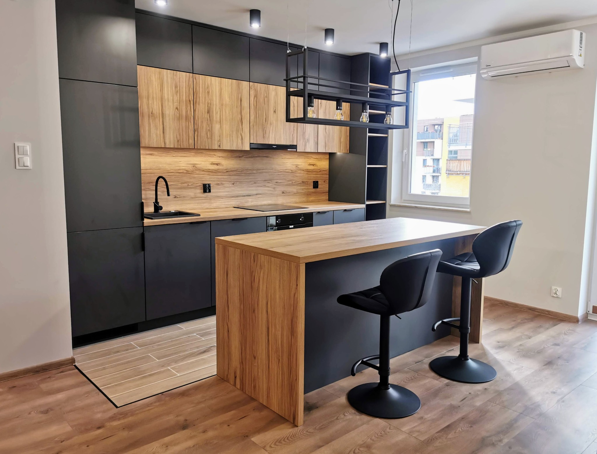 Nowoczesna kuchnia z czarnymi szafkami i drewnianymi frontami, wyspa z dwoma czarnymi hokerami, oświetlenie punktowe i wiszące lampy w industrialnym stylu, widok na budynek za oknem.
