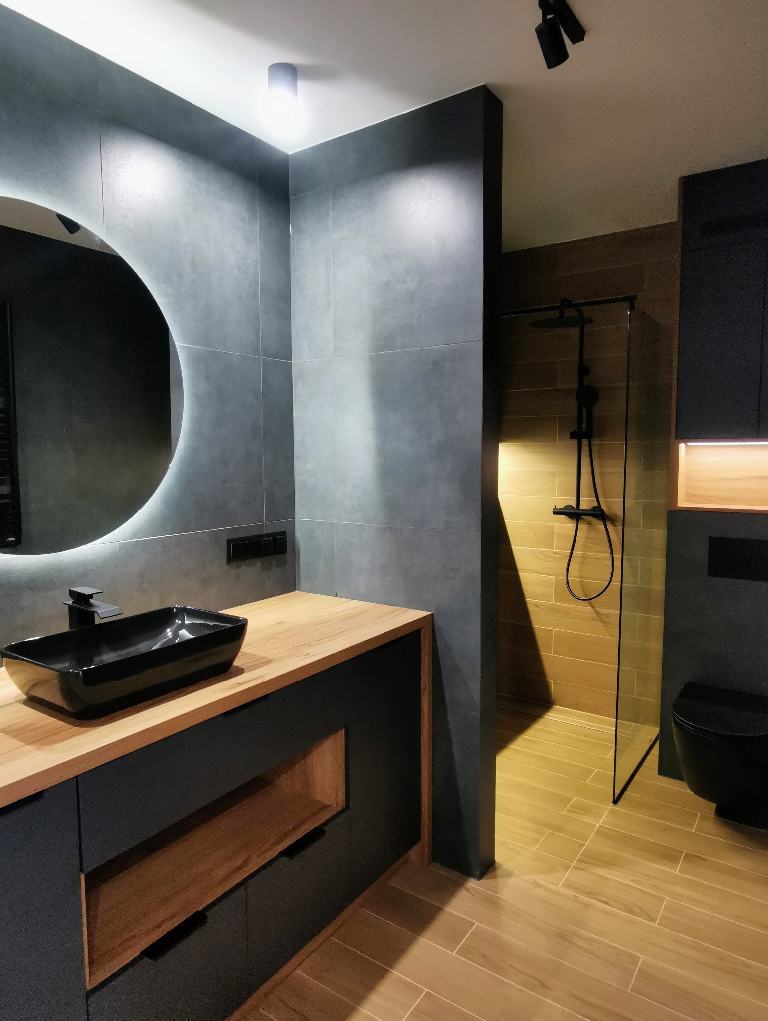 Nowoczesna łazienka z szarymi płytkami, okrągłym lustrem z podświetleniem LED, czarną umywalką nablatową i drewnianym blatem, oraz kabiną prysznicową z czarną armaturą.