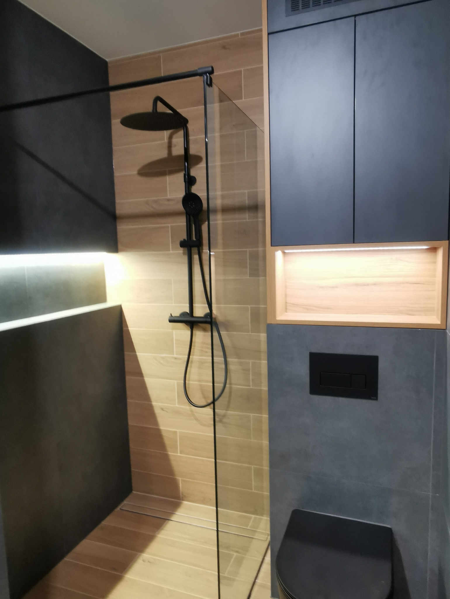 Nowoczesna łazienka z kabiną prysznicową typu walk-in z czarną armaturą, drewnopodobnymi płytkami na ścianie prysznica i szarą zabudową meblową z podświetlaną wnęką.