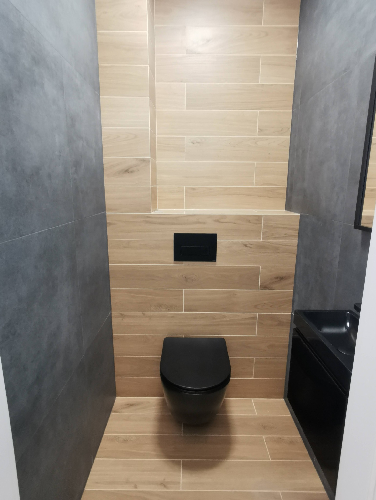 Nowoczesna toaleta w odcieniach szarości i beżu z podwieszaną miską WC w kolorze czarnym i czarnym przyciskiem spłukującym na ścianie wyłożonej płytkami imitującymi drewno.