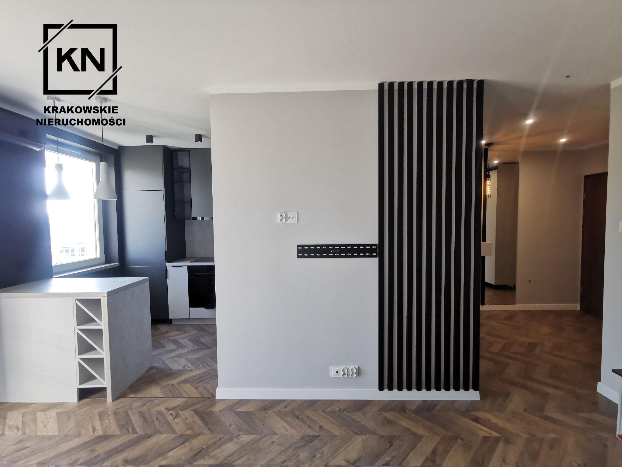 Nowoczesne wnętrze mieszkania z parkietem w jodełkę, białym aneksem kuchennym z półkami na wino, ścianą z pionowymi czarnymi lamelami i uchwytem do telewizora.