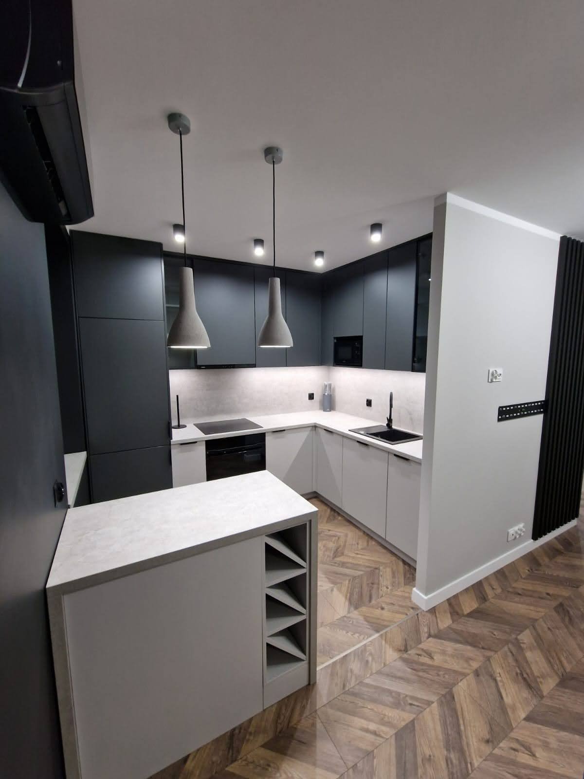 Nowoczesna kuchnia w odcieniach szarości z drewnianą podłogą w jodełkę, wiszące lampy stożkowe i wyspa z półkami na wino.