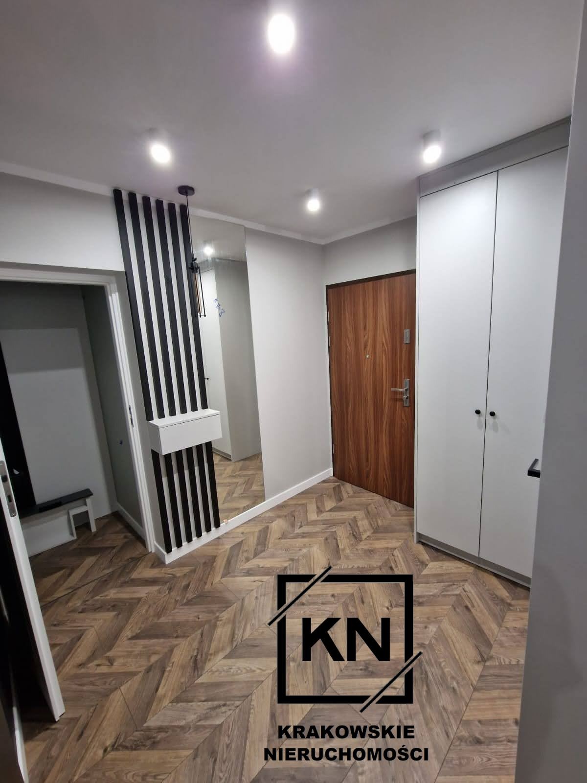 Nowoczesny przedpokój z parkietem w jodełkę, lustrem z dekoracyjnymi czarnymi listwami i białą szafą wnękową.