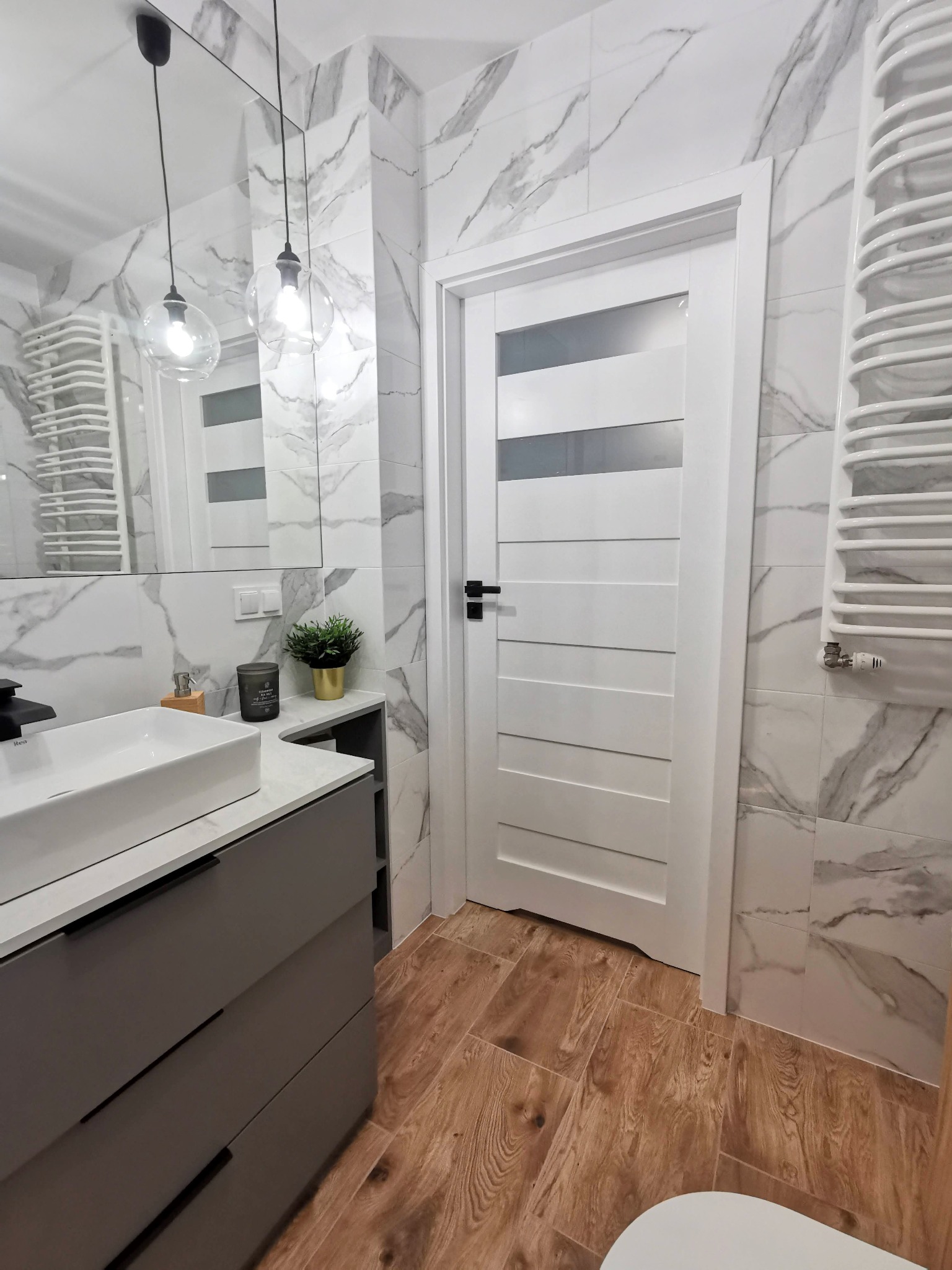 Nowoczesna łazienka z marmurowymi płytkami, drewnopodobną podłogą, szarą szafką pod umywalką i białą, panelową, szklaną  drzwi.