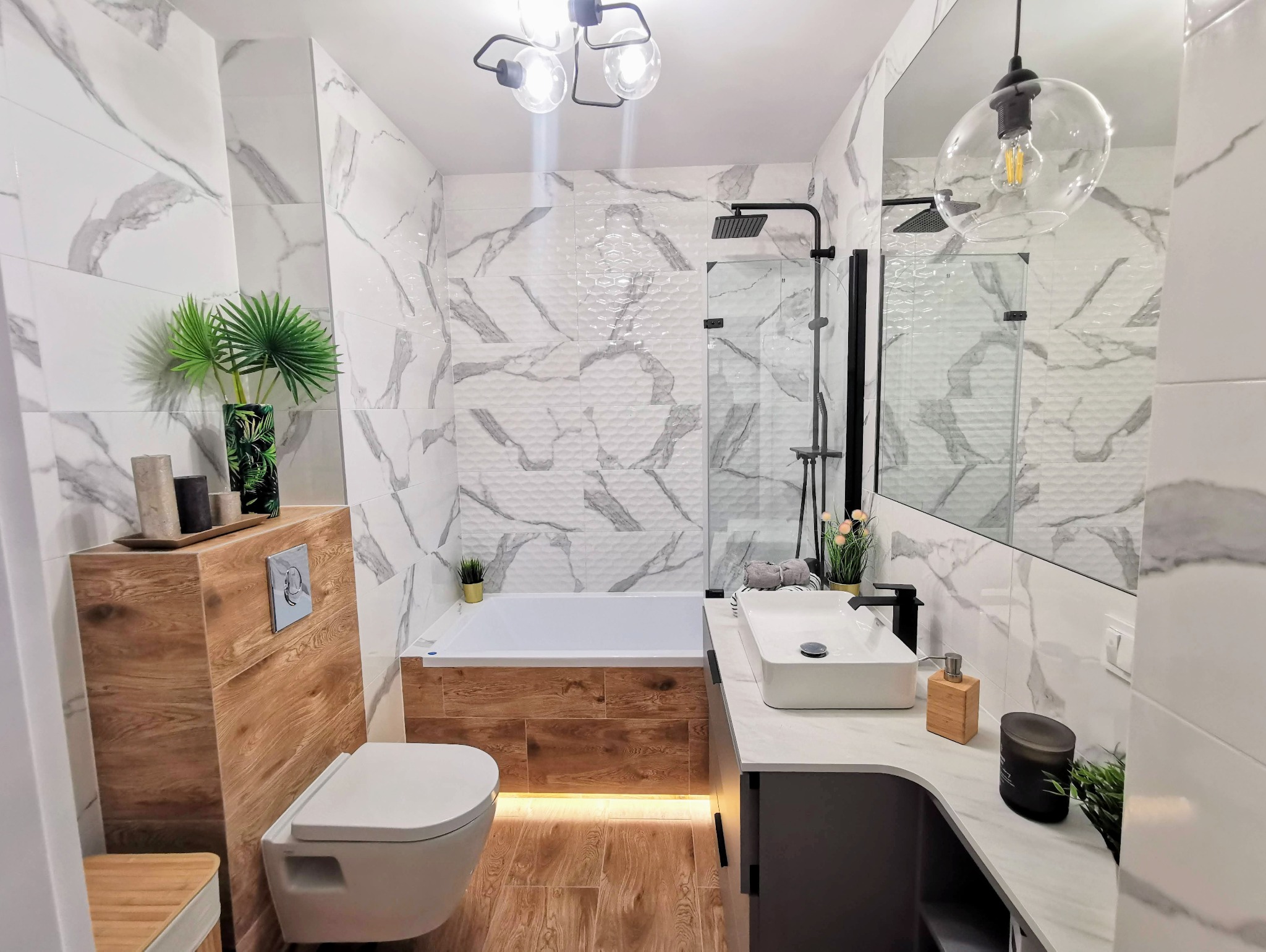 Nowoczesna łazienka z białymi płytkami imitującymi marmur, drewnianymi akcentami na obudowie wanny i zabudowie WC, czarną armaturą, oraz oświetleniem dekoracyjnym.